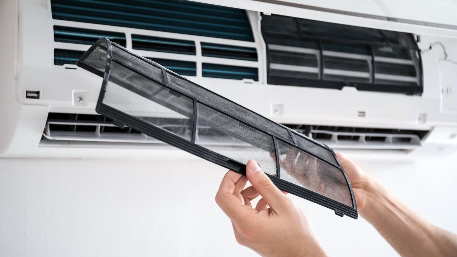 Hands removing a rectangular air conditioner filter from an indoor wall-mounted unit in preparation for cleaning or replacement.
